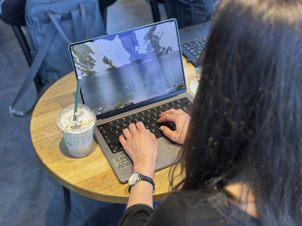A person typing on a laptop at a small round wooden table, with an iced drink beside it