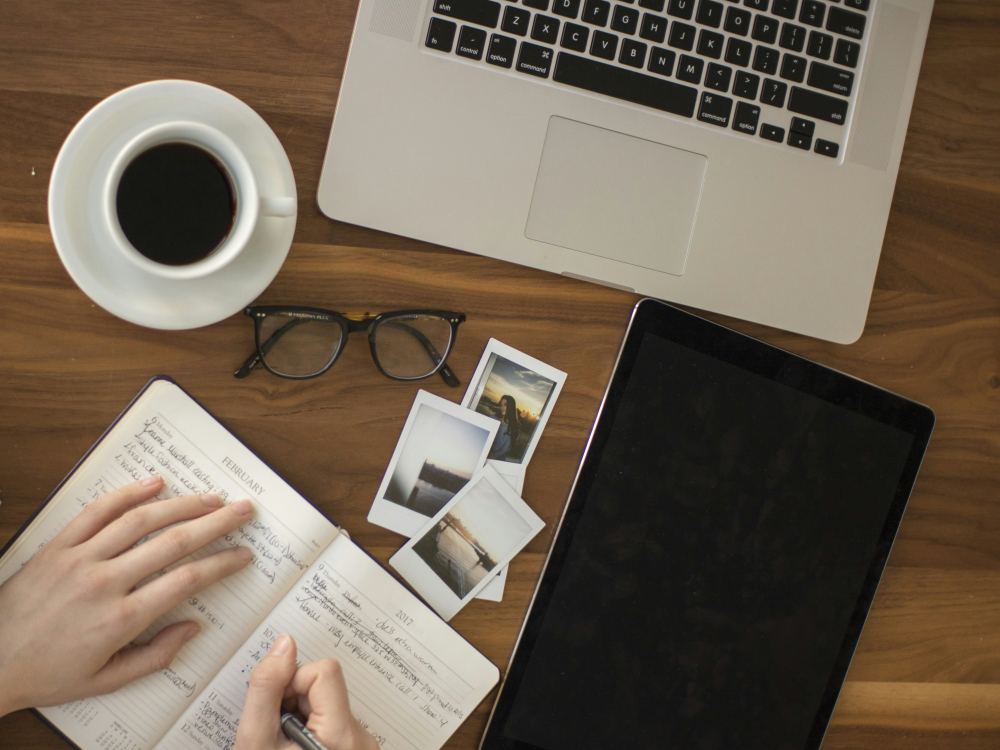 A person writing in a notebook at a wooden desk, with a laptop and a tablet nearby